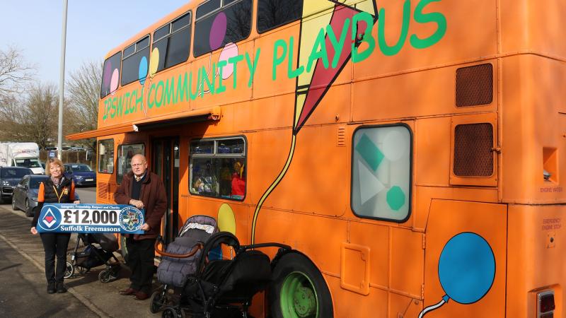 two people stand outside a bus holding a banner that says '£12,000 Suffolk Freemasons'. The Bus is a bright orange double decker bus with Ipswitch Community Playbus written in green writing. There are balloons and a kit painted onto the side. two people stand outside a bus holding a banner that says '£12,000 Suffolk Freemasons'. The Bus is a bright orange double decker bus with Ipswitch Community Playbus written in green writing. There are balloons and a kit painted onto the side.